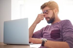 Homem de óculos com a mão no rosto diante do notebook, com olho cansado